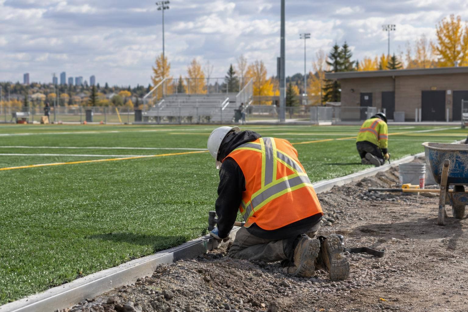 Two newly completed artificial turf fields with crews finishing field-edge work, lighting poles, spectator seating and washroom facilities visible in the same frame.