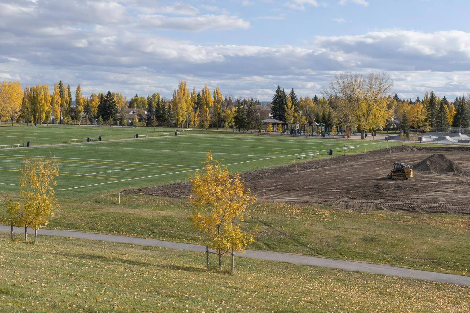 A documentary view across Fonda Park showing the completed flag football fields beside the area being prepared for the third field, with the existing playground and skate slab visible in the background.